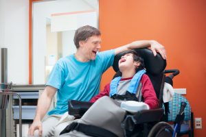 Father talking with disabled biracial son sitting in wheelchair while waiting in doctor's office, laughing together.